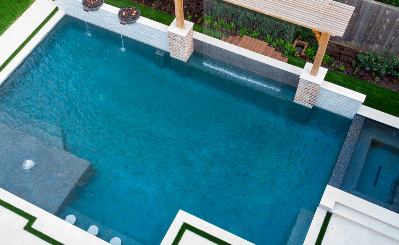 Aerial view of a custom geometric swimming pool with integrated spa, shear descent water features, fire bowls, and tanning ledge.