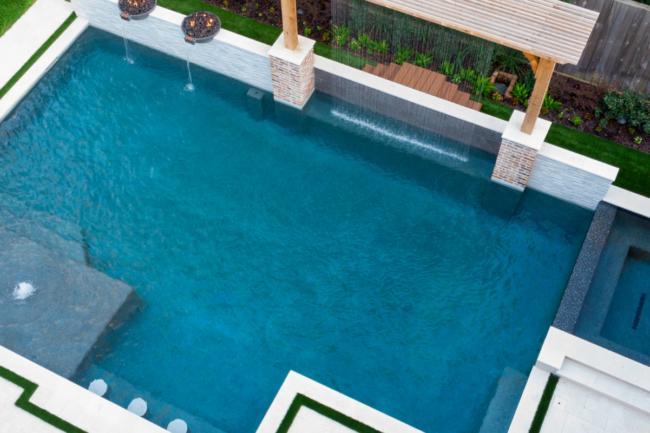Aerial view of a custom geometric swimming pool with integrated spa, shear descent water features, fire bowls, and tanning ledge.