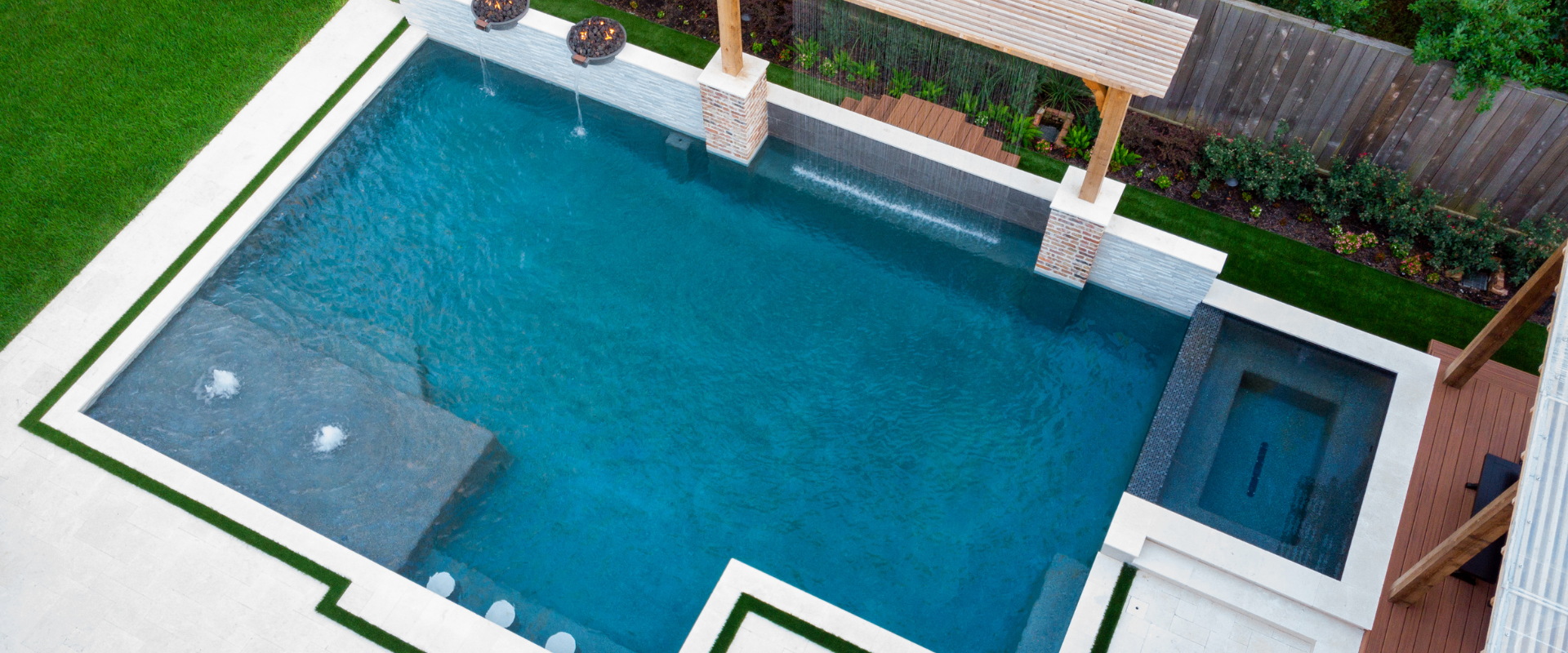 Aerial view of a custom geometric swimming pool with integrated spa, shear descent water features, fire bowls, and tanning ledge.