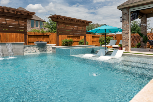 Custom backyard pool featuring a shallow tanning ledge with white loungers, umbrella, sheer descent water features on a stone wall, and a covered patio dining area.