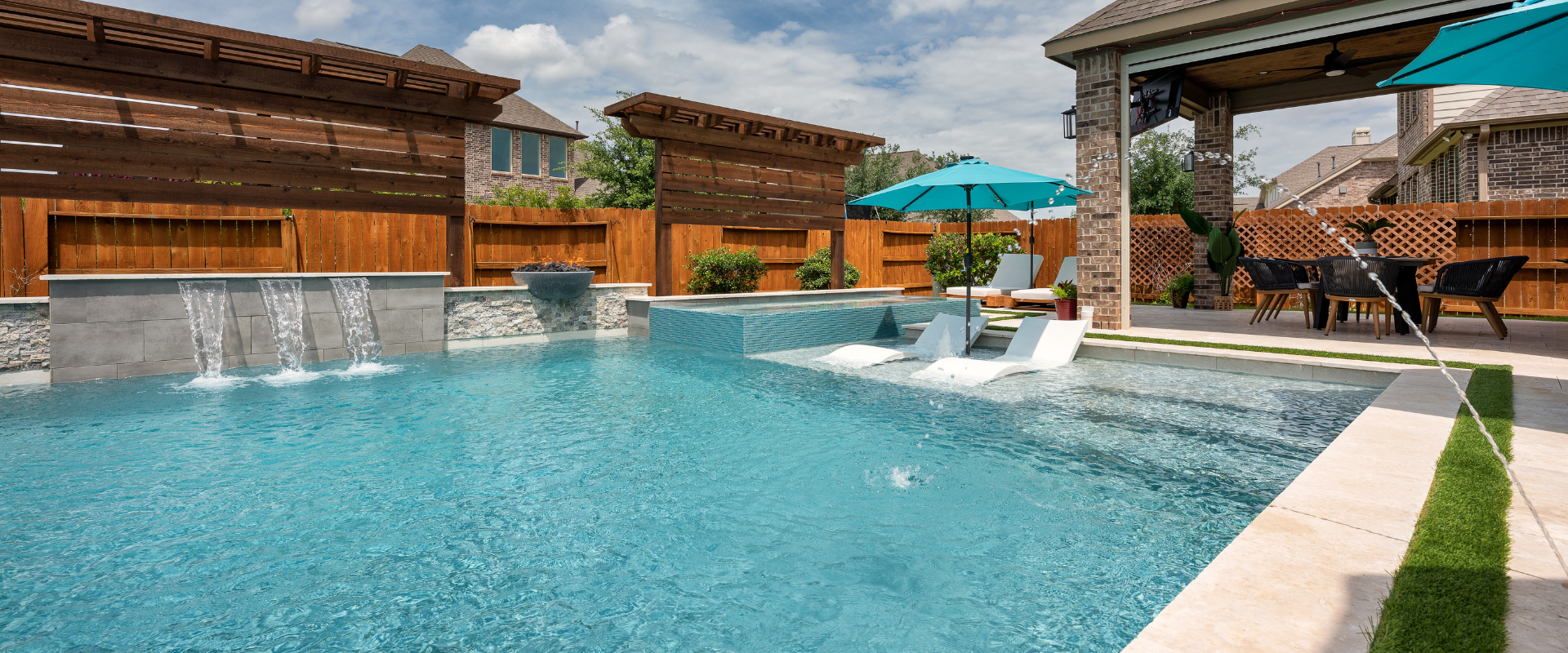 Custom backyard pool featuring a shallow tanning ledge with white loungers, umbrella, sheer descent water features on a stone wall, and a covered patio dining area.