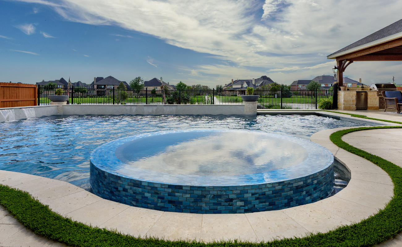 High angle view of a luxury custom pool with an integrated circular blue tile spa, water feature wall, paver deck with artificial turf strips, and covered patio kitchen.