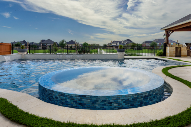 High angle view of a luxury custom pool with an integrated circular blue tile spa, water feature wall, paver deck with artificial turf strips, and covered patio kitchen.