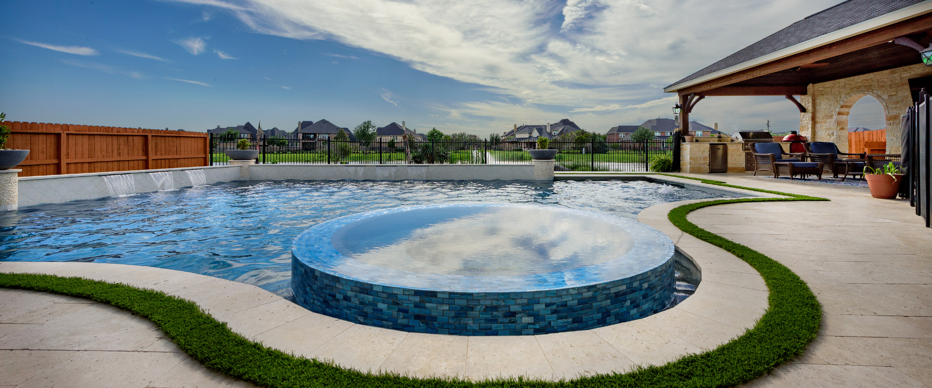 High angle view of a luxury custom pool with an integrated circular blue tile spa, water feature wall, paver deck with artificial turf strips, and covered patio kitchen.