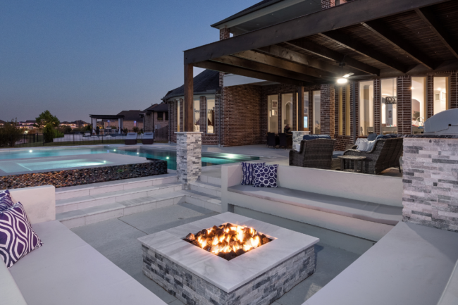 Twilight view of a sunken fire pit lounge with built-in seating, an adjacent raised spa, and a covered outdoor living patio with kitchen and lighting.
