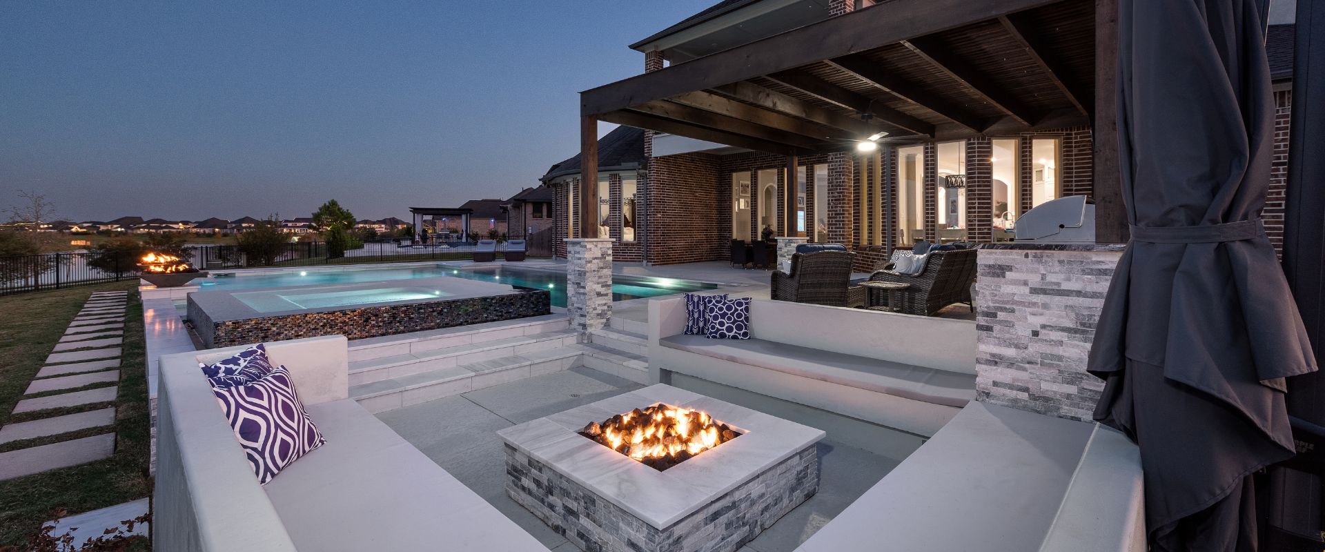 Twilight view of a sunken fire pit lounge with built-in seating, an adjacent raised spa, and a covered outdoor living patio with kitchen and lighting.